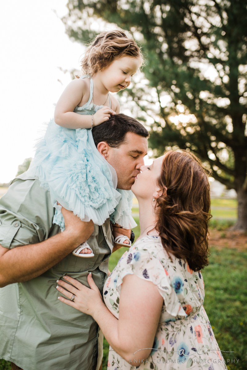 child sitting on dads shoulders looking down while he kisses her mother photographed by Anika Colombo Photography