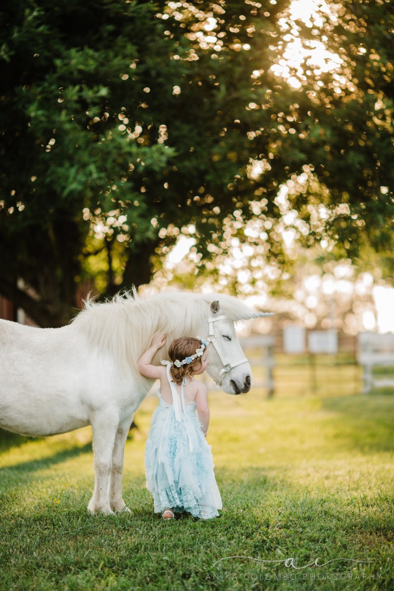 child standing in a field under a tree facing away from the camera while petting a sleepy unicorn