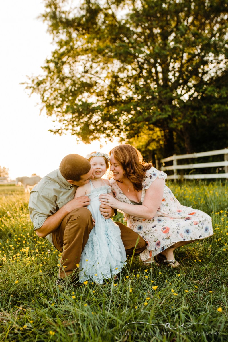 toddler staring into he camera as parents tickle and laugh at her in a field