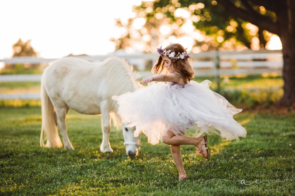 little girl wearing big tulle dress and flower crown twirling in the foreground with a white unicorn in the background, photo by Anika Colombo Photography
