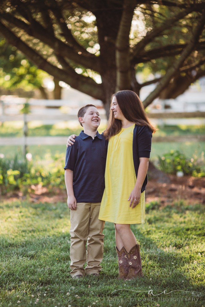 Siblings standing in front of a tree with arms around each other looking at each other and laughing