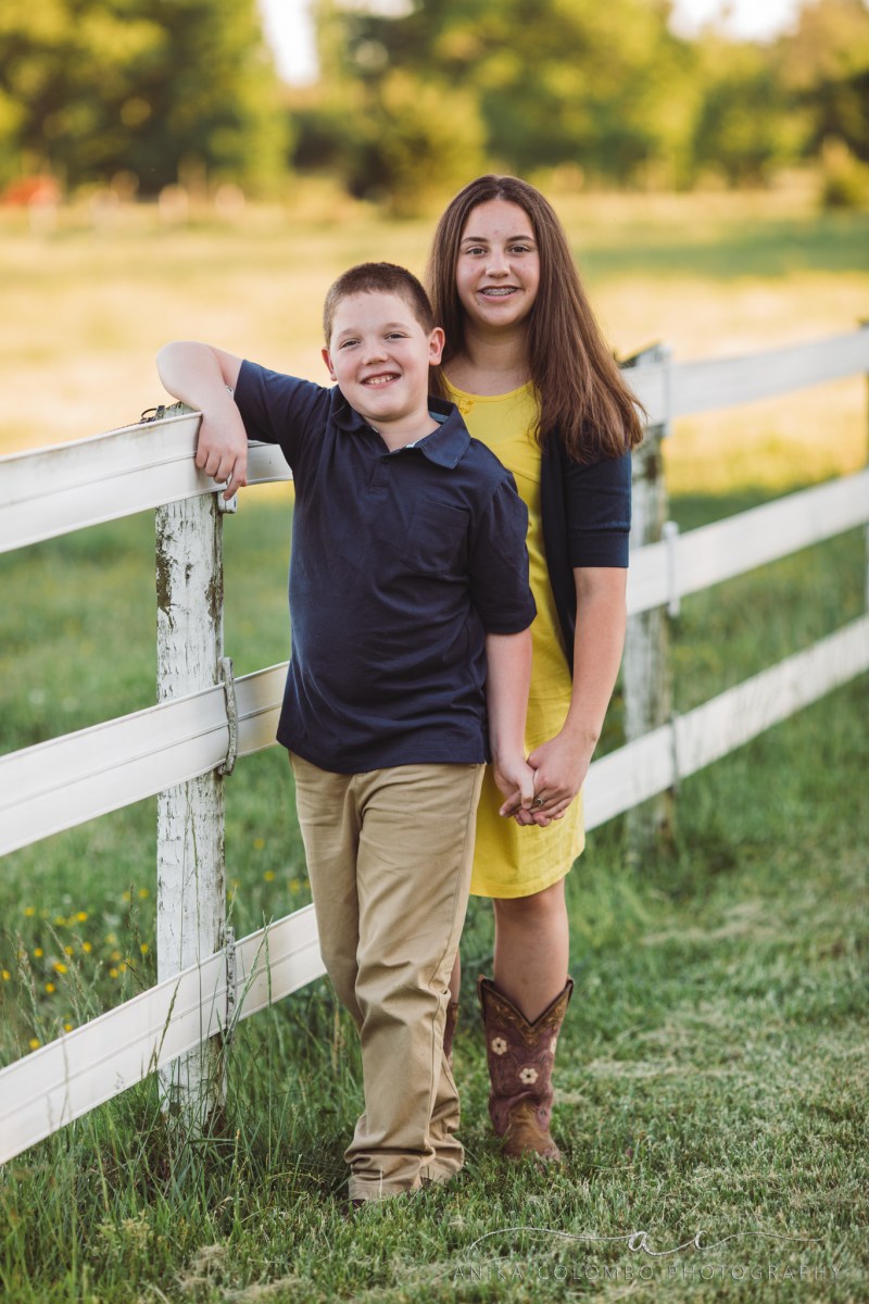 brother and sister standing at a fence to a pasture, sister is standing behind her younger brother, arms on the fence, other hands holding each other