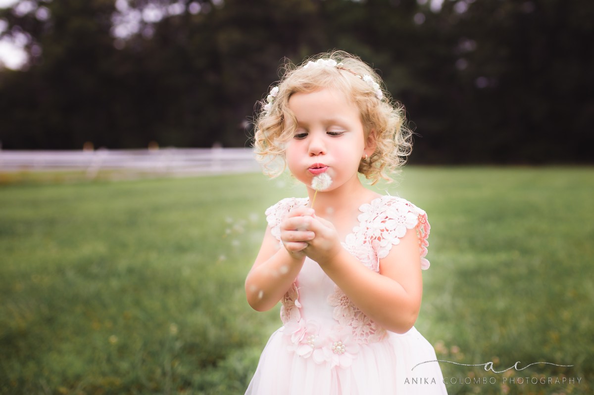 little girl wearing a pink dress and floral crown blowing a dandelion