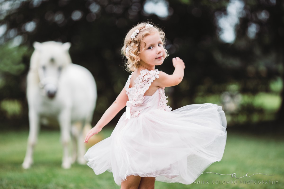 little girl wearing pink dress and floral crown twirling with a white unicorn in the background