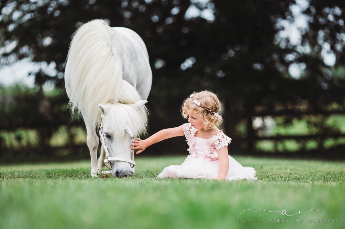 little girl wearing pink dress and floral crown sitting in the grass petting a white unicorn's head