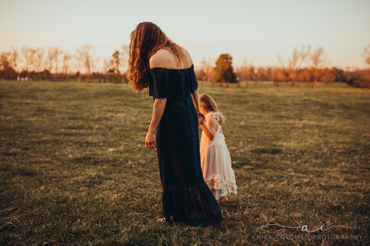 mother walks hand in hand with young daughter in a field at sunset