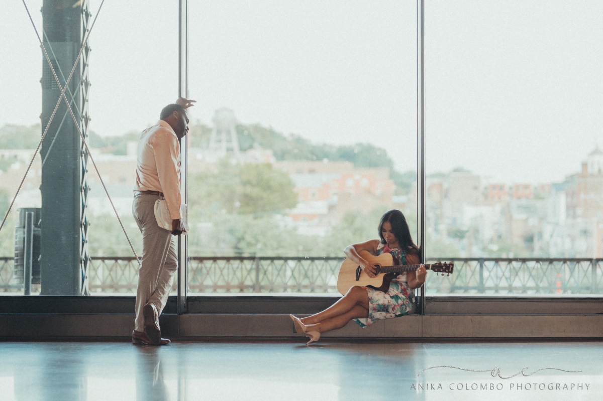 woman playing a guitar in mainstreet station in front of big windows overlooking the city while a man looks on leaning on the window holding music sheets